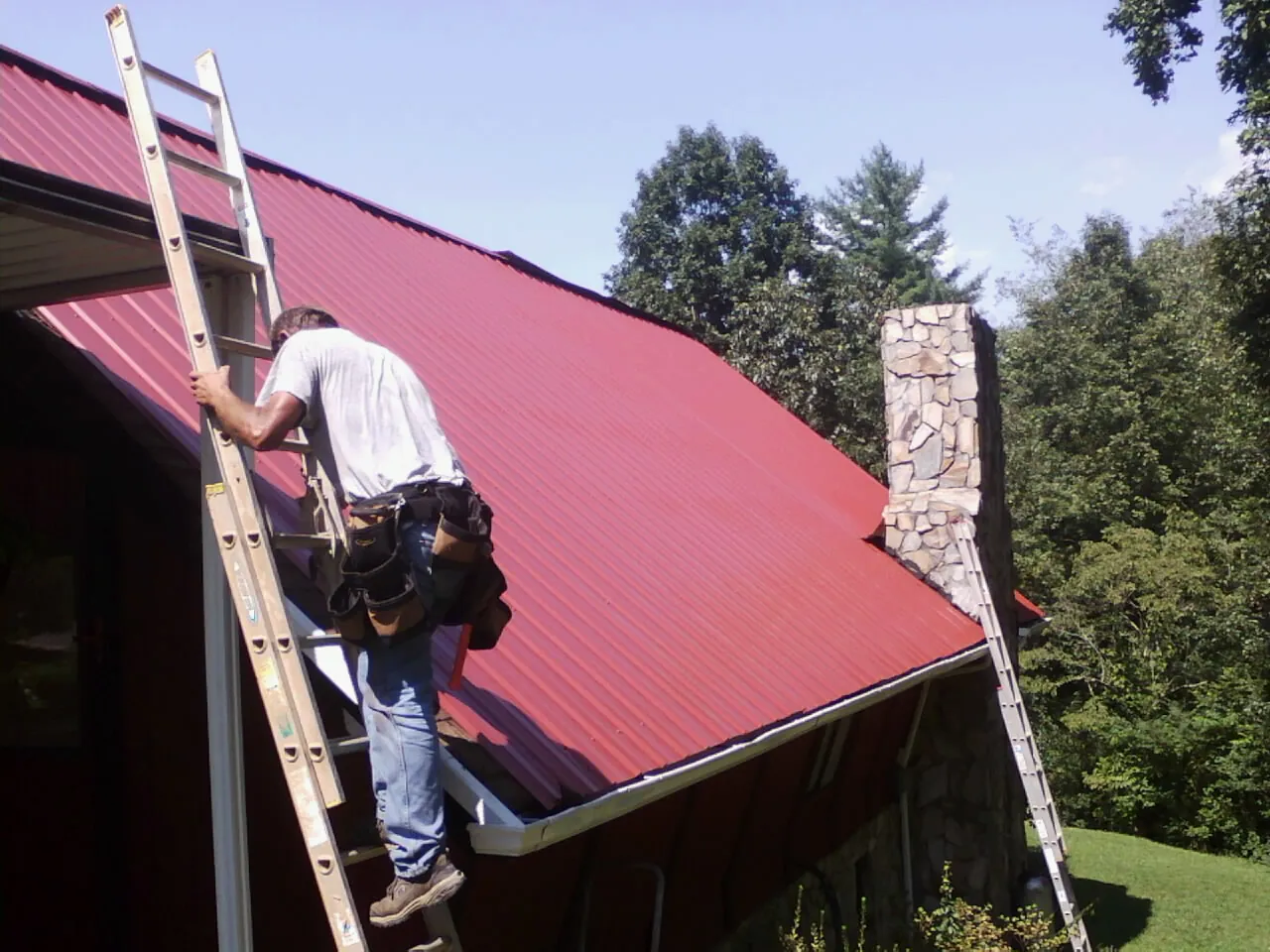 Roofer on a ladder beside a red metal roof and stone chimney — Soffit & Fascia Repair by Roofing Brothers in Byers